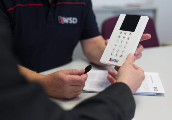 ChatGPT: Auf dem Bild sieht man zwei Personen, die gemeinsam ein Bedienteil einer Alarmanlage betrachten. Eine Person hält das weiße Gerät mit Tastatur und Display in der Hand, die andere hält einen schwarzen Transponder. Beide sitzen an einem Tisch, auf dem auch Unterlagen mit dem WSD-Logo liegen. Das Bild vermittelt eine professionelle Beratungssituation im Sicherheitsbereich.